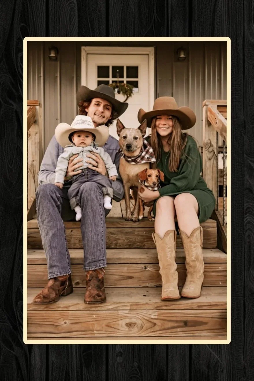 Family photo of a man, woman, two dogs, and a baby sitting on wooden porch steps in front of a house with a door decorated with a wreath and siding.