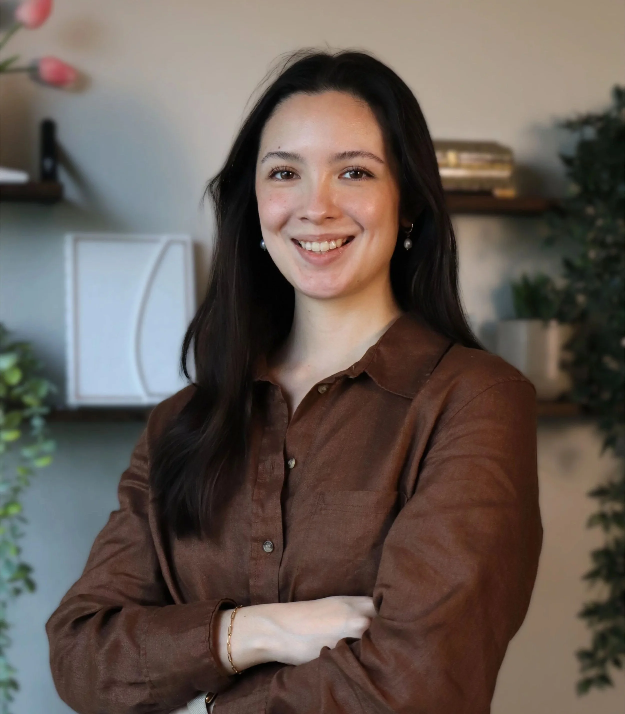 A woman with long dark hair smiling, wearing a brown button-up shirt, standing with her arms crossed in a cozy indoor setting with plants and shelves in the background.