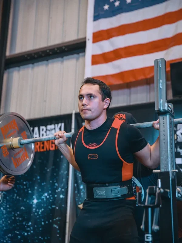 A man preparing to lift a barbell during a powerlifting competition, with an American flag hanging in the background.