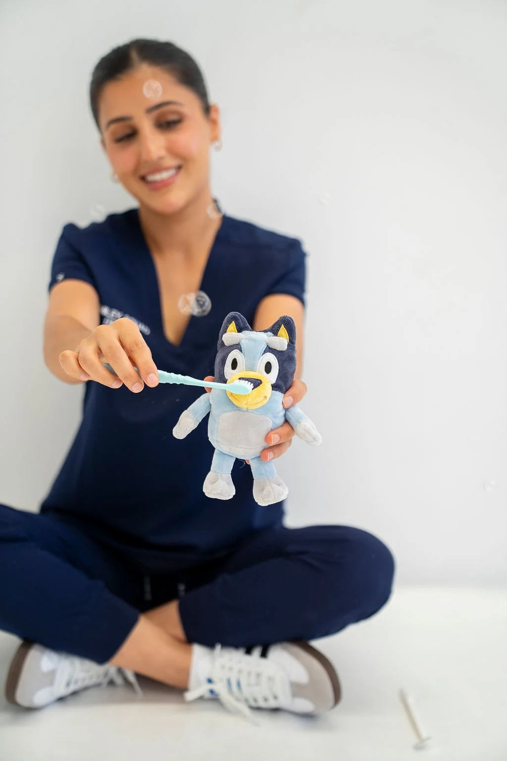 A woman sitting cross-legged on the floor, smiling, holding a plush toy of a cartoon character, and brushing its teeth with a small toothbrush.