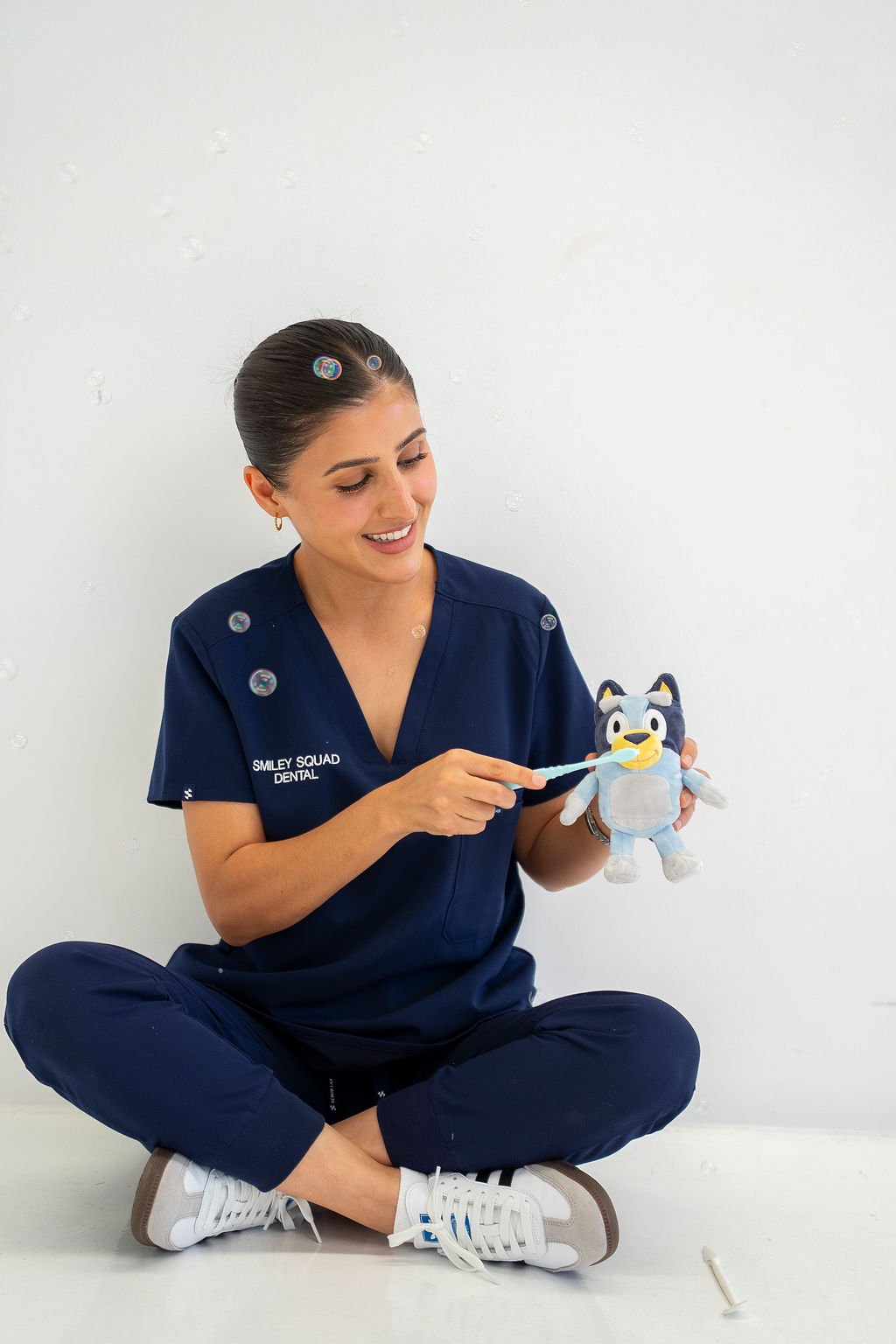 A woman in navy dental scrubs sitting cross-legged against a white wall, holding a plush toy of a blue cartoon character, and brushing the toy's teeth with a toothbrush, with soap bubbles floating around.