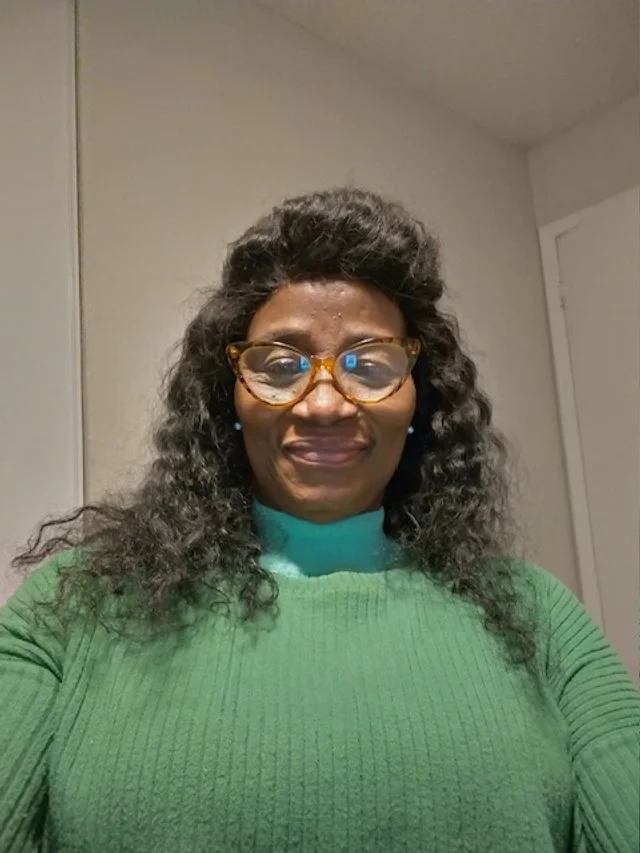 A woman with medium-dark skin, curly hair, and glasses, wearing a green sweater, taking a selfie indoors in front of a plain wall.