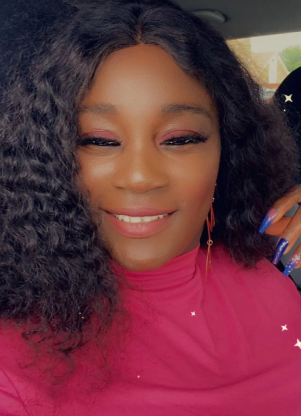 Close-up of a woman smiling with curly black hair, wearing pink lipstick, pink eyeshadow, a pink top, and long blue decorated nails.