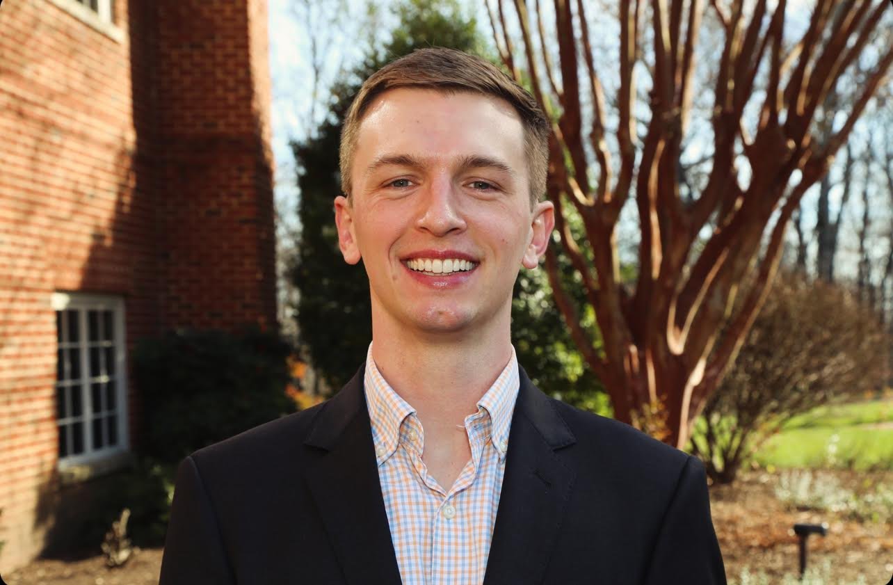 Young man smiling outdoors in front of a brick building and trees.
