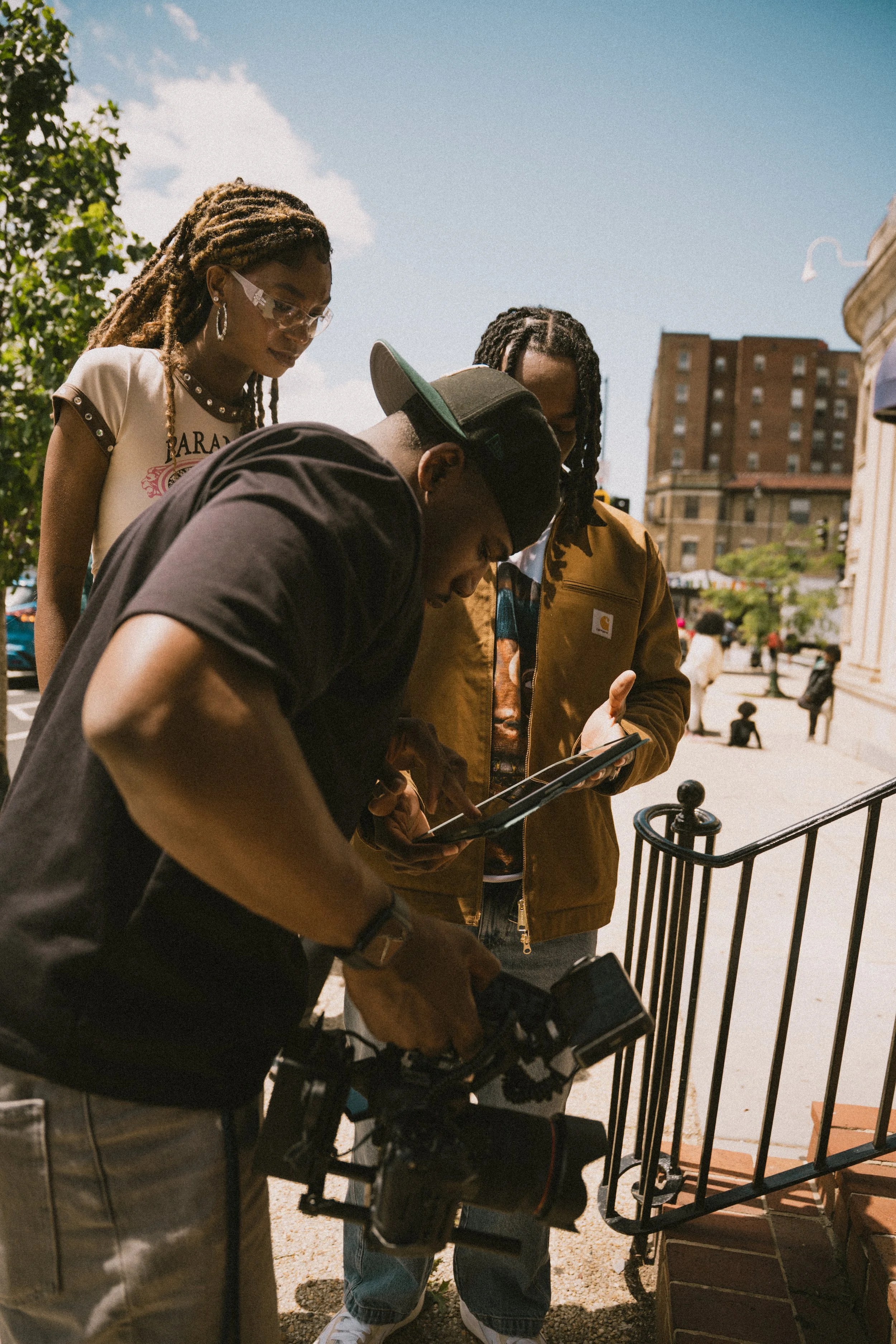 Four young people gathered outdoors around a camera and a tablet, looking at the devices, with a building and people in the background on a sunny day.