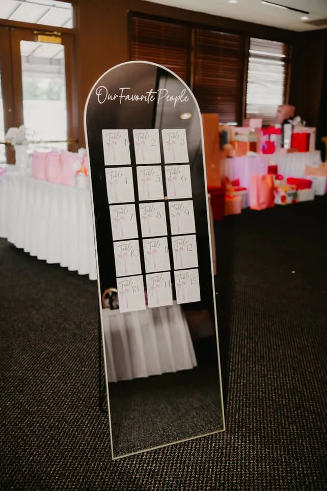 A standing mirror with a list of table numbers for an event, labeled "Our Favorite People" at the top, with gift bags and presents in the background.