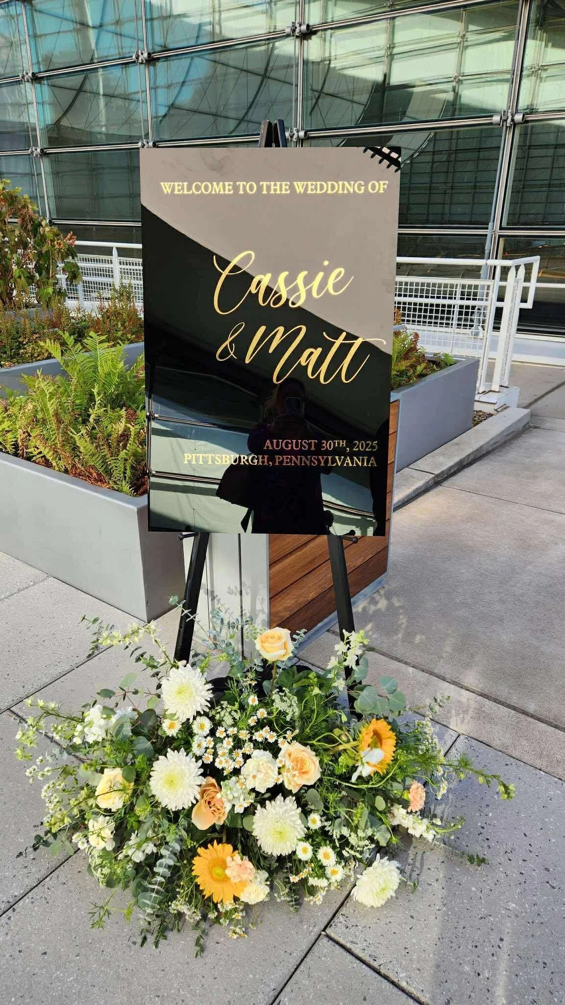 A wedding welcome sign with floral arrangements at the base, reading 'Welcome to the wedding of Cassie & Matt, August 30th, 2025, Pittsburgh, Pennsylvania', placed outside a modern building.
