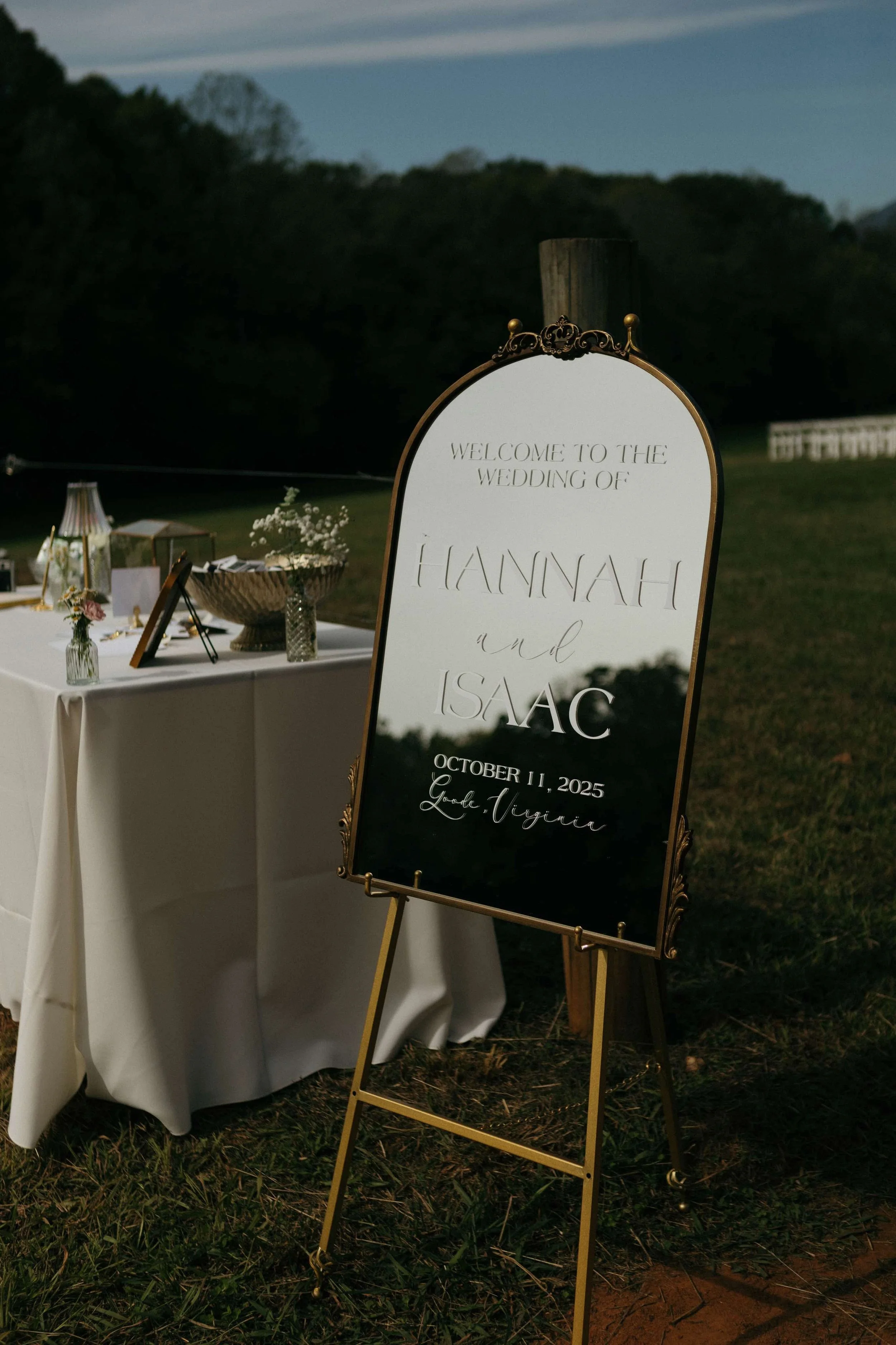 Wedding welcome sign with gold frame on grass, featuring elegant script text with names Hannah and Isaac, date October 11, 2025, and location Lord Virginia, next to a decorated table at dusk.