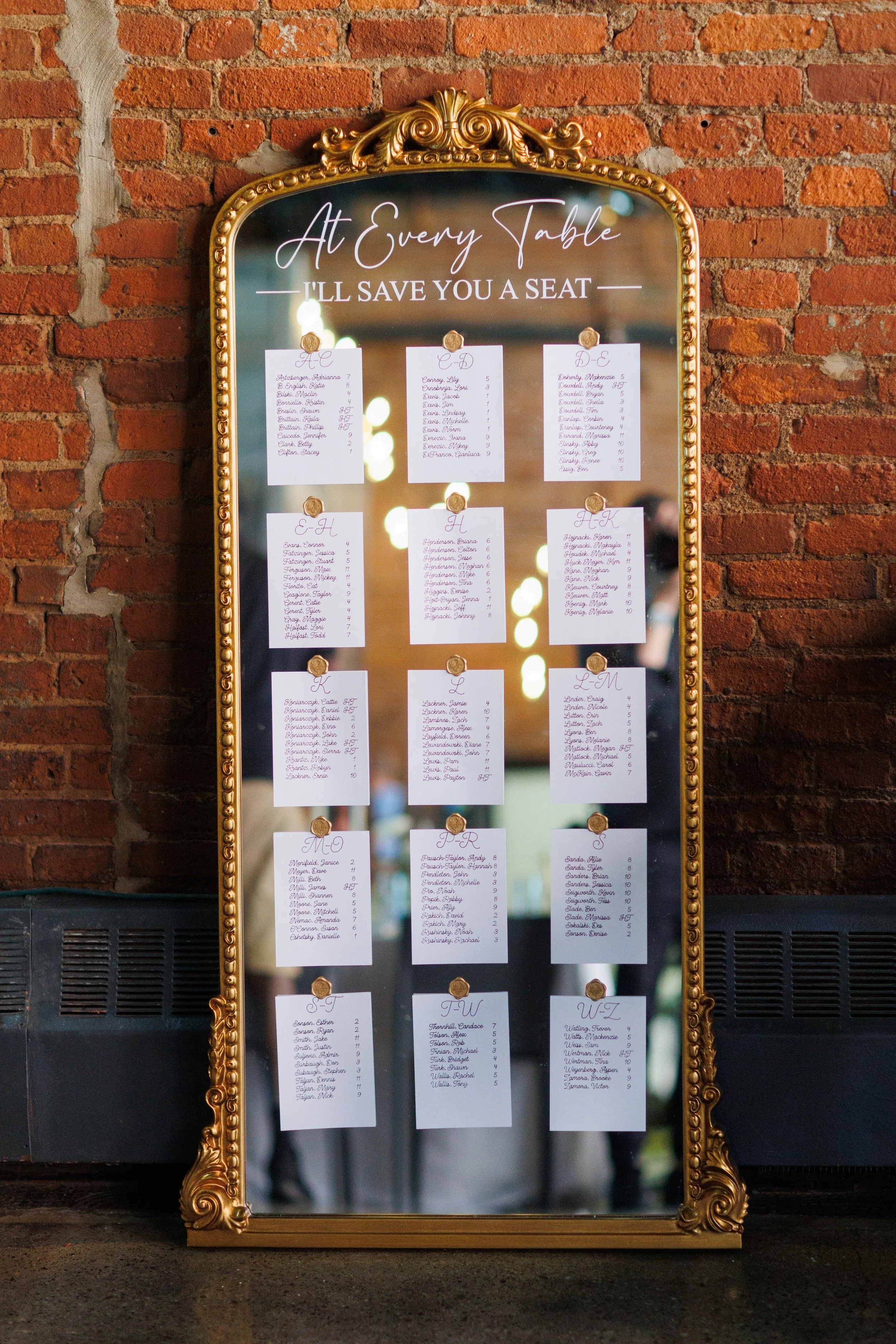 A gold-framed mirror with a seating chart for a wedding reception, displaying tables named alphabetically from A to W, with guest names listed underneath each table.