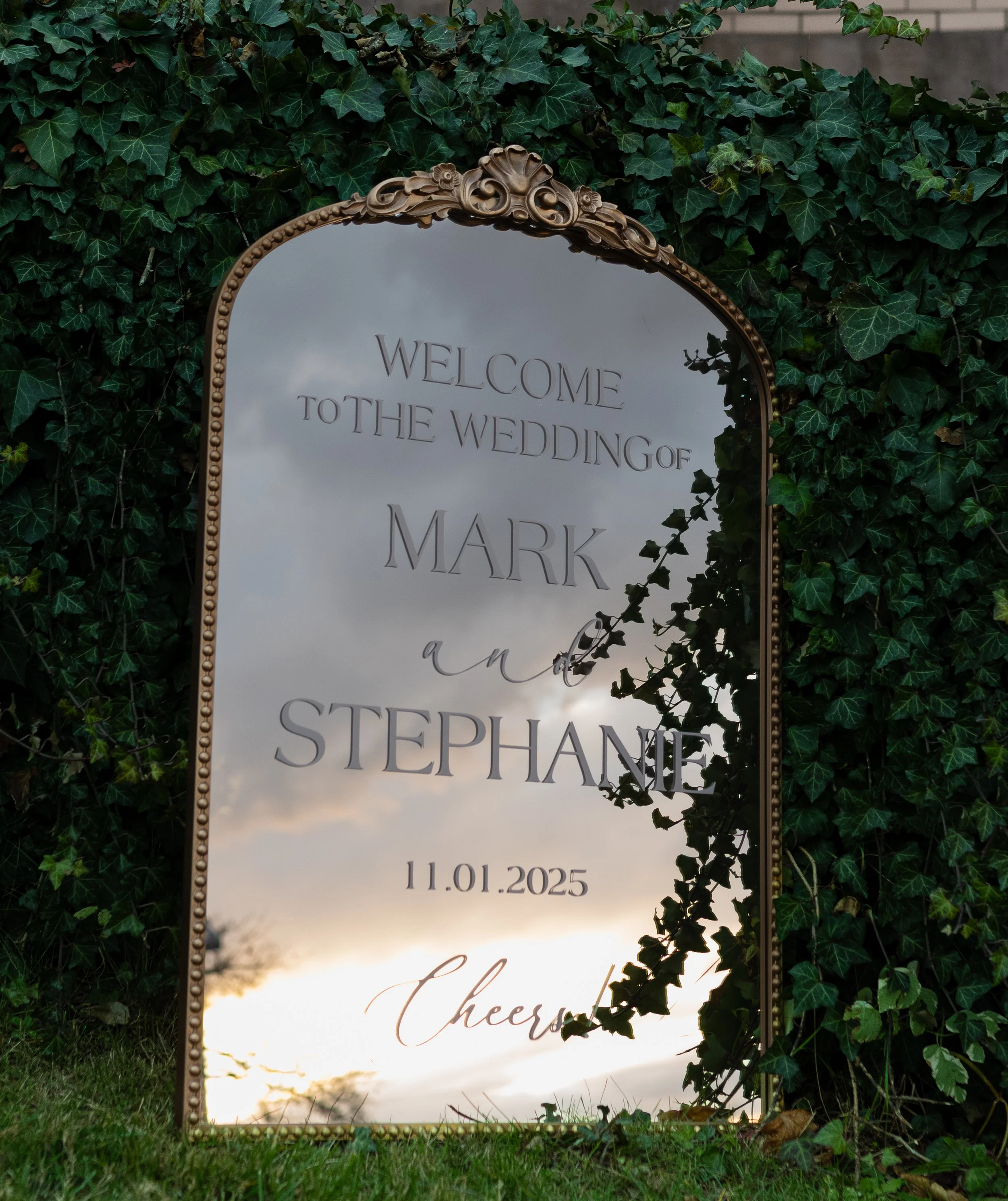 Decorative wedding mirror sign with gold frame reading "Welcome to the wedding of Mark and Stephanie, 11.01.2025," surrounded by green ivy, outdoors with cloudy sky reflection.