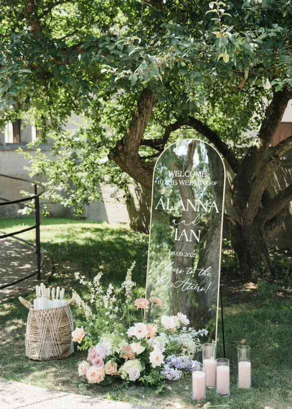 A wedding sign on a mirror with floral arrangements and candles in a garden setting.