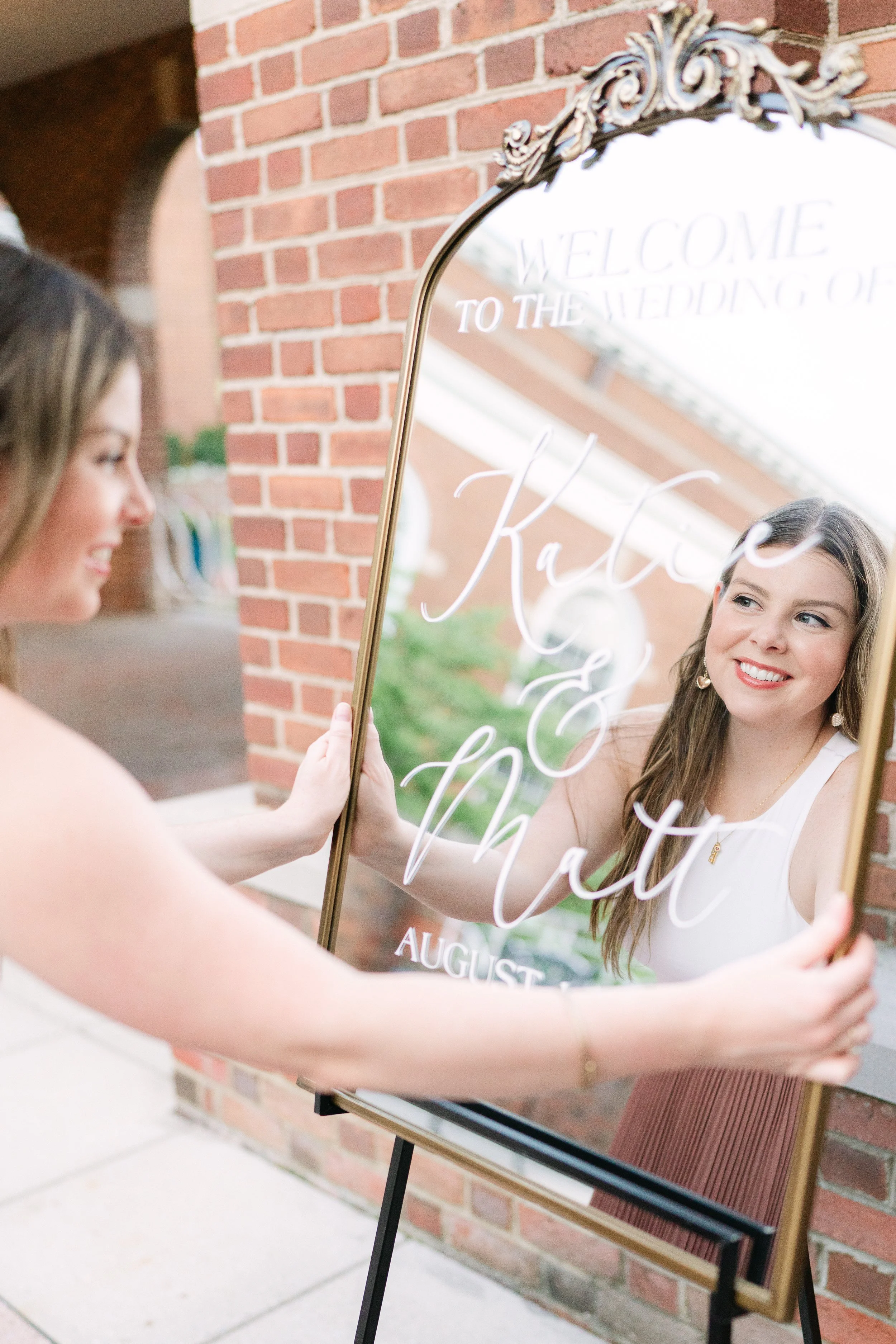 A woman is holding a decorative mirror with wedding information written on it, including the names Katie and Matt, and the wedding date in August. The mirror reflects her smiling face, and she is standing outside near a brick wall.