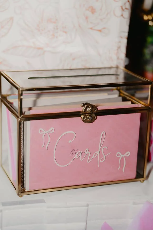 Glass display box labeled 'Cards' with pink background and white bows, containing greeting cards, on a white surface.