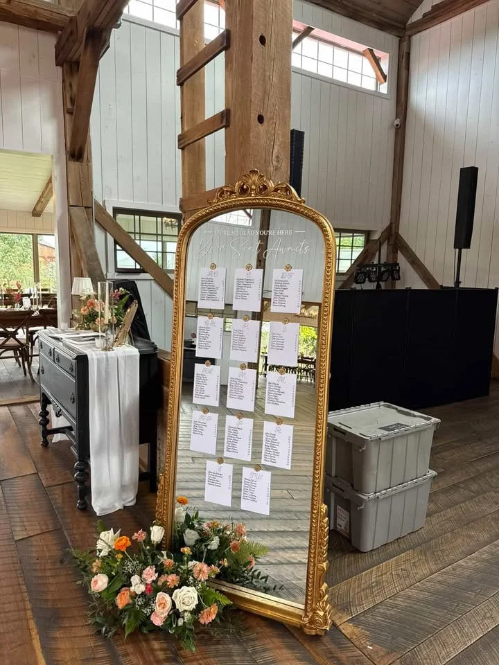 A large, ornate, gold-framed mirror with wedding seating arrangements pinned to it, decorated with a flower arrangement at the base. The mirror reflects the wooden interior of a rustic venue with high ceilings and natural light.