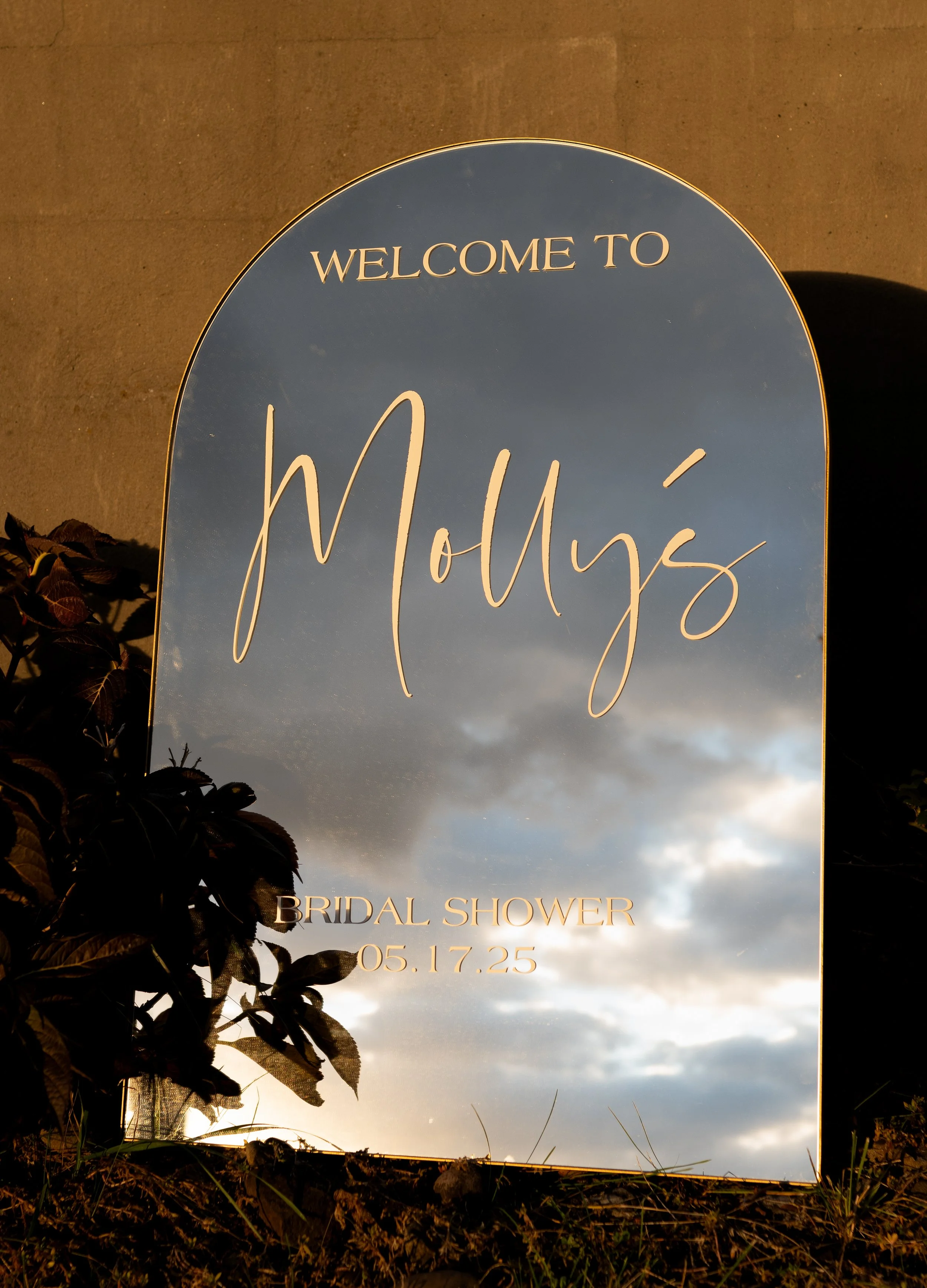 Mirror-like welcome sign for Molly's bridal shower, with cloudy sky reflection, surrounded by dark leaves and grass at the base.