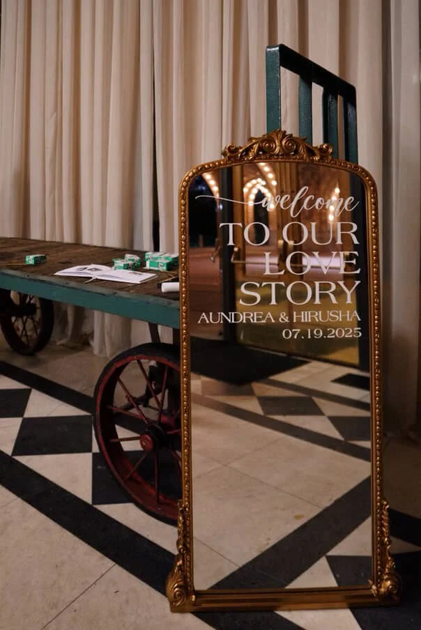 Decorative mirror with wedding message for Andrea and Hirusha's wedding on July 19, 2025, reflecting string lights in the background.