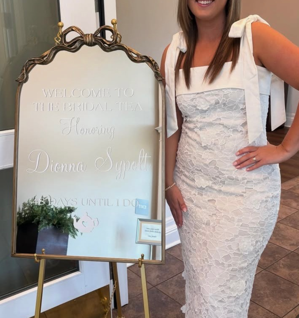 A woman in a white lace dress standing next to a sign with elegant writing that reads "Welcome to the Bridal Tea honoring Dianna Supatt" and a countdown of 48 days until the event.