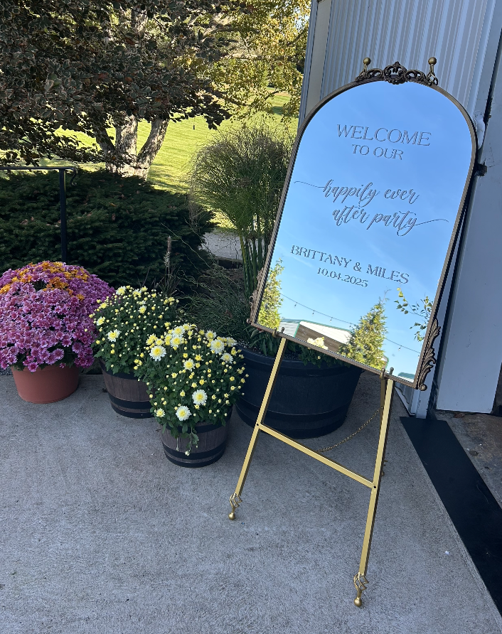 A large mirror with ornate frame and stand placed outside near potted flowers, welcoming guests to a celebration for Brittany and Miles on October 4, 2025.