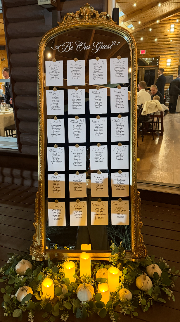 A decorative wedding seating chart with white papers pinned to a large ornate gold-framed mirror. The chart is titled 'Be Our Guest' and lists tables numbered one to sixteen with names of guests underneath each number. The mirror is decorated with ca