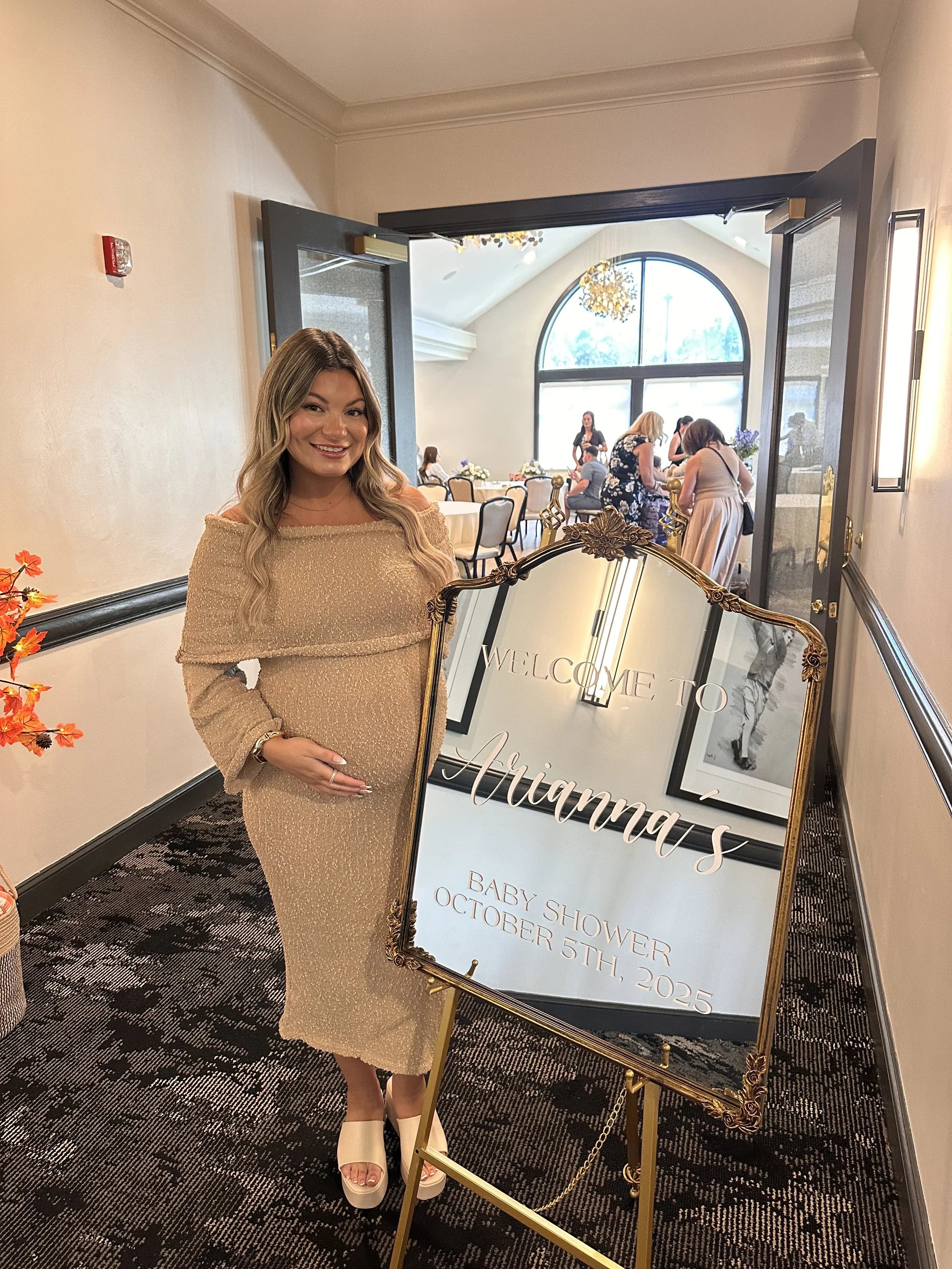 A pregnant woman in a beige dress smiling and holding her belly, standing next to a welcome sign for a baby shower on October 5th, 2025, in a decorated event space.