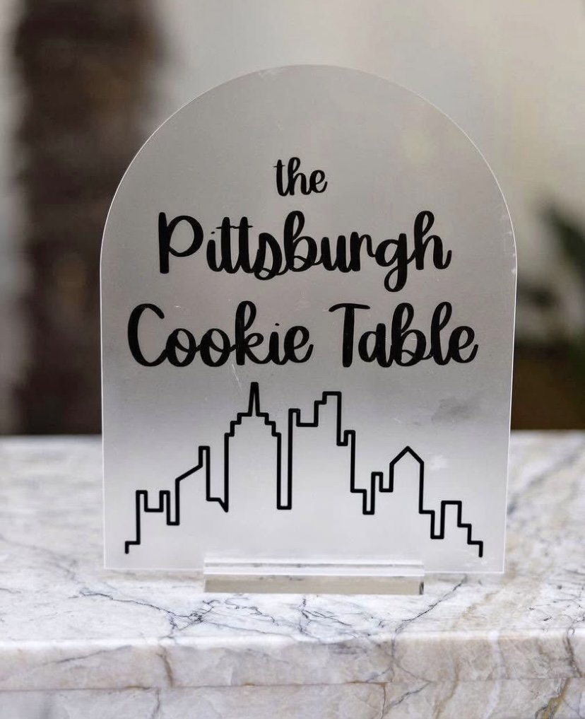 A clear acrylic sign with black lettering that reads "the Pittsburgh Cookie Table" and a black outline of the Pittsburgh skyline, placed on a marble surface.