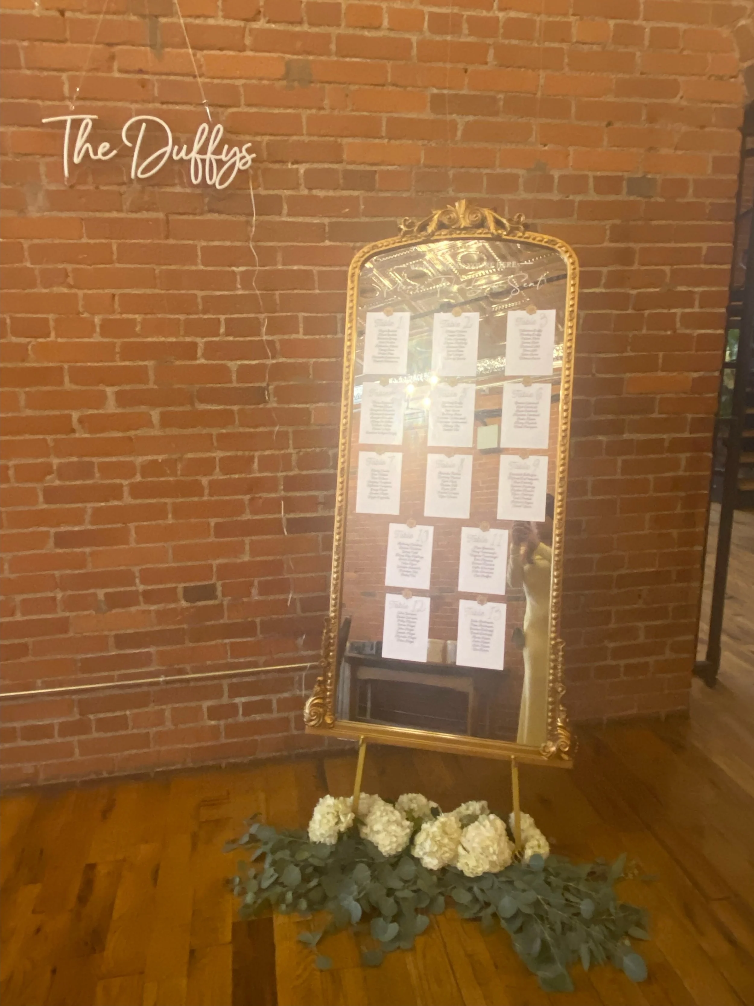 A large vintage gold-framed mirror leaning against an exposed brick wall, decorated with white flowers and green foliage at its base. The mirror reflects the interior ceiling and lights. To the left, a white illuminated sign reads "The Duffy's".