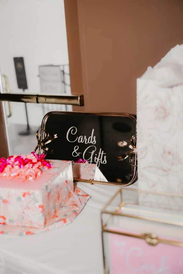 Decorative card and gift display with a black sign reading 'Cards & Gifts' in cursive font, pink flowers on wrapped presents, and other gift items on a table.