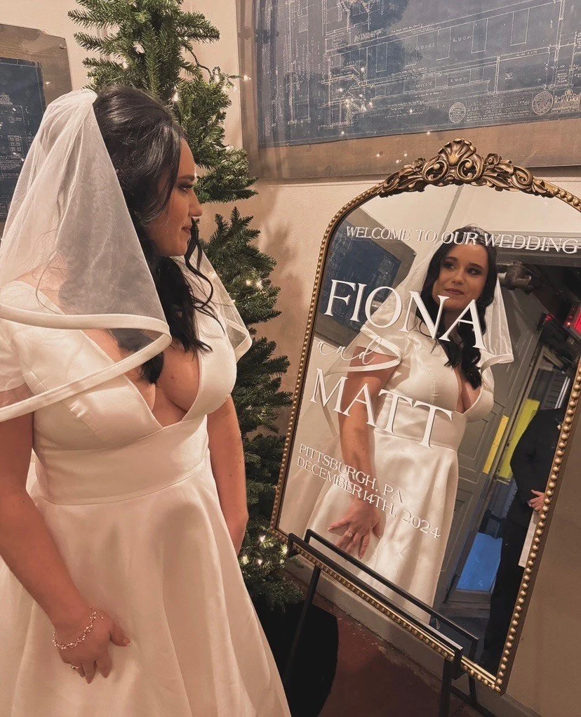 A woman in a white wedding dress with a veil is looking at her reflection in a mirror. The mirror has text that says 'Welcome to our wedding, Fiona and Matt, Pittsburgh, PA, December 14th, 2024.' There is a decorated Christmas tree beside her and a framed picture of a blueprint on the wall.