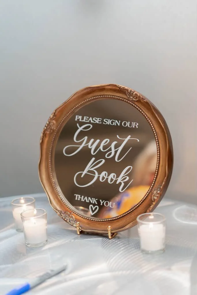 Decorative mirror with a wooden frame reading, 'Please sign our guest book, thank you,' on a table with candles.