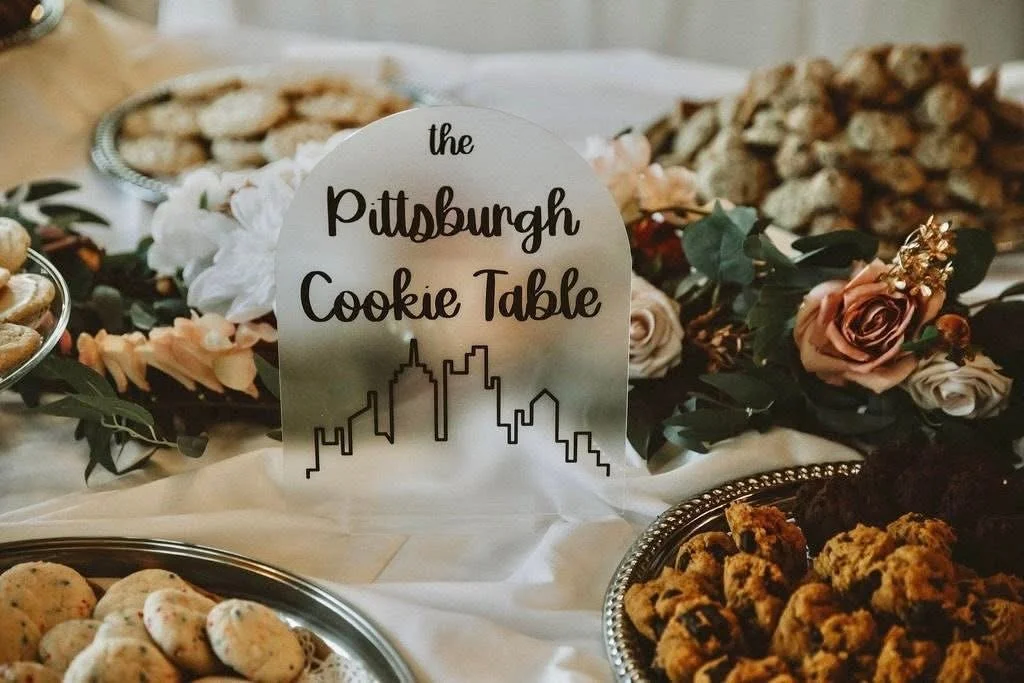 Decorative table with cookies, a floral arrangement, and a sign that reads 'the Pittsburgh Cookie Table' with a city skyline outline.