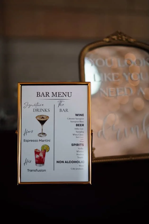 Framed bar menu with sections for signature drinks, beer, wine, spirits, and non-alcoholic beverages, featuring images of a cocktail and a Transfusion drink.