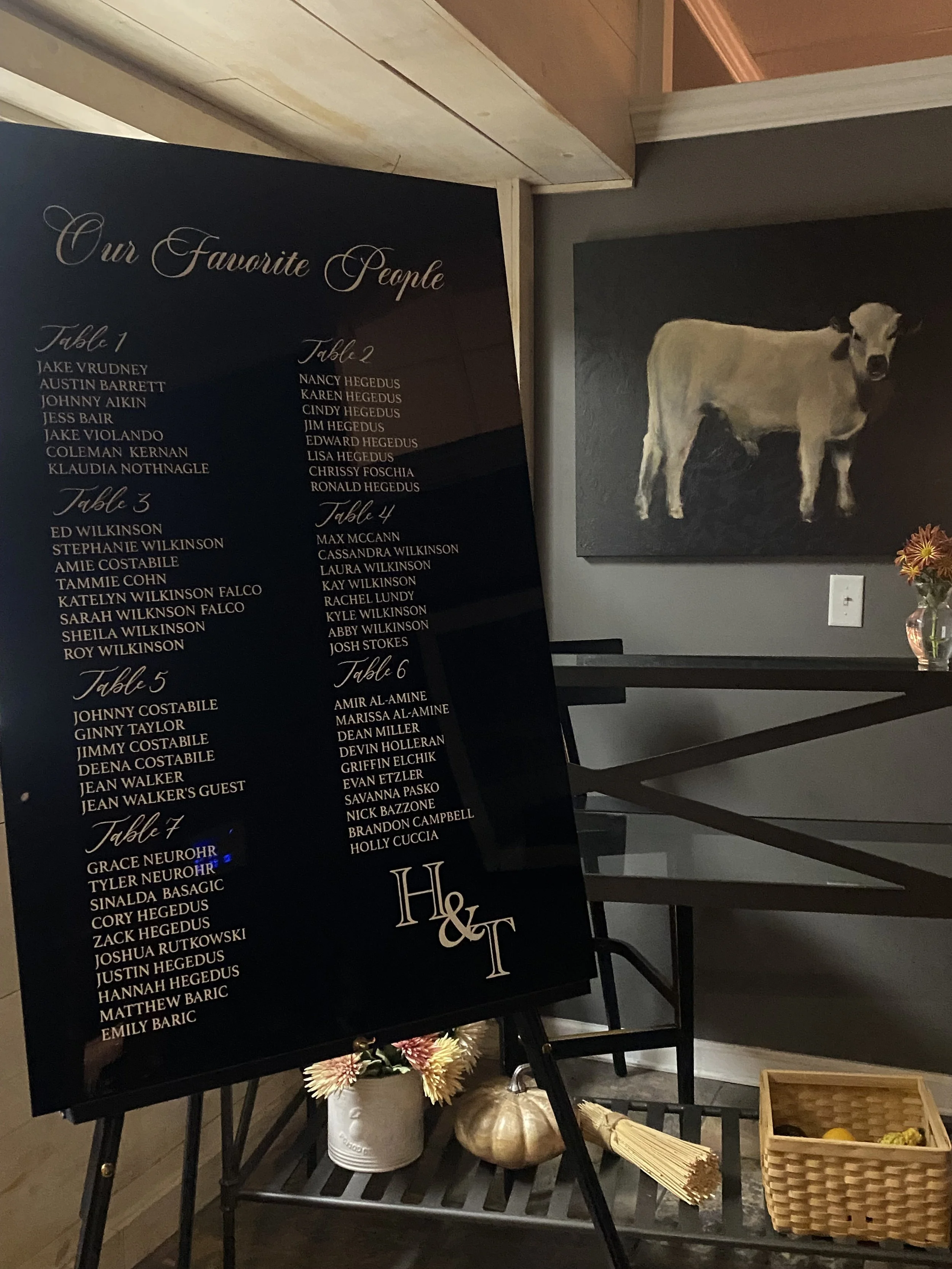 A black wedding seating chart with gold and white writing listing guest names for seven tables, and the initials 'H & T' at the bottom. The chart is placed on an easel, beside a small table with flowers, a pumpkin, and other decorative items. There i