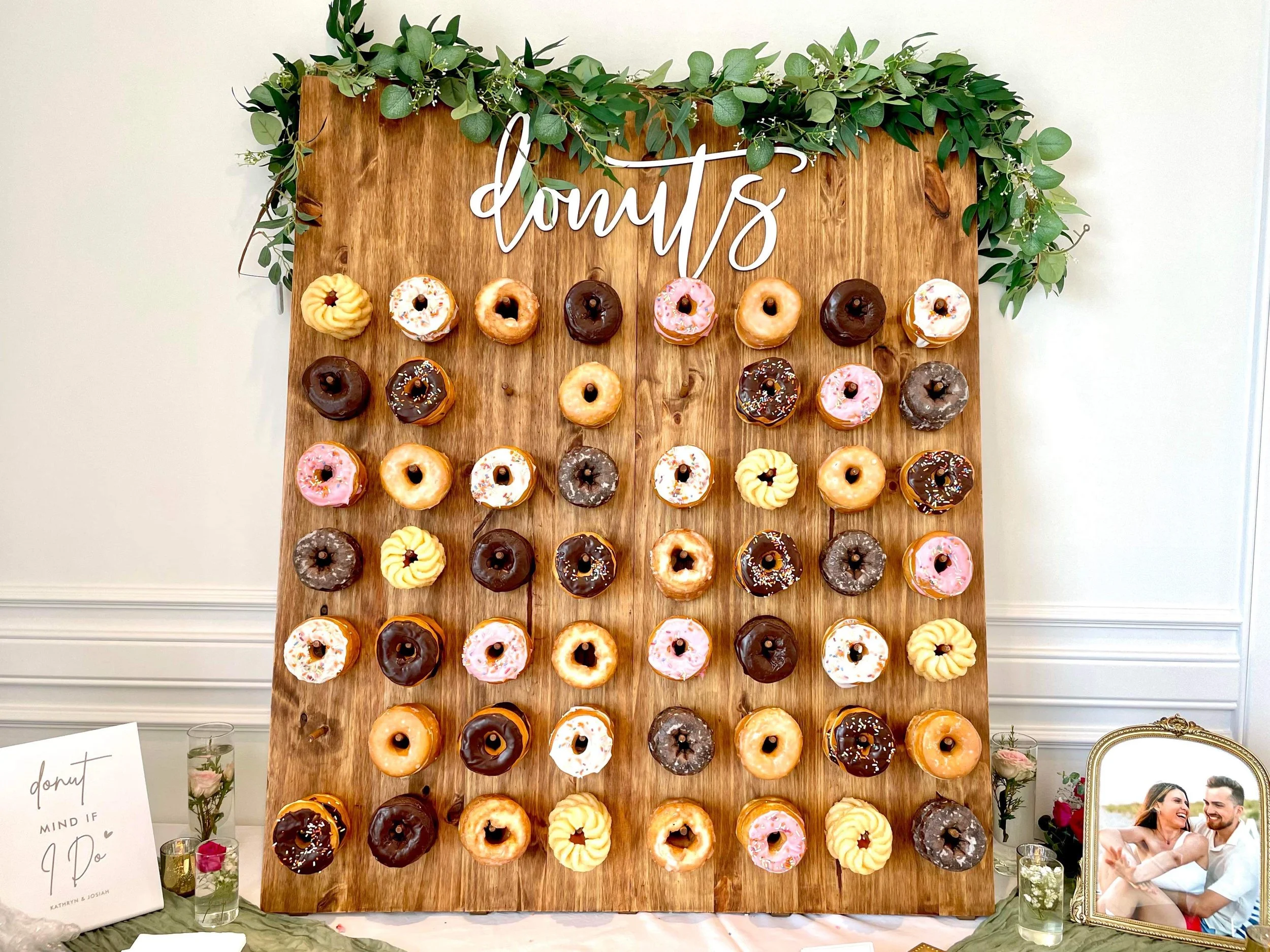 A large wooden board decorated with a variety of colorful doughnuts arranged in rows, topped with sprinkles and icing, and a green leafy garland at the top. The word 'doughnuts' is written in cursive at the top of the board. To the left, there is a s