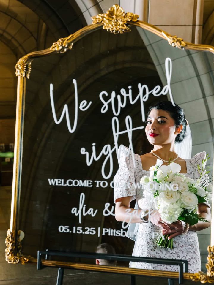 Bride in white lace dress holding a bouquet of white flowers in front of a decorative mirror with a wedding message.