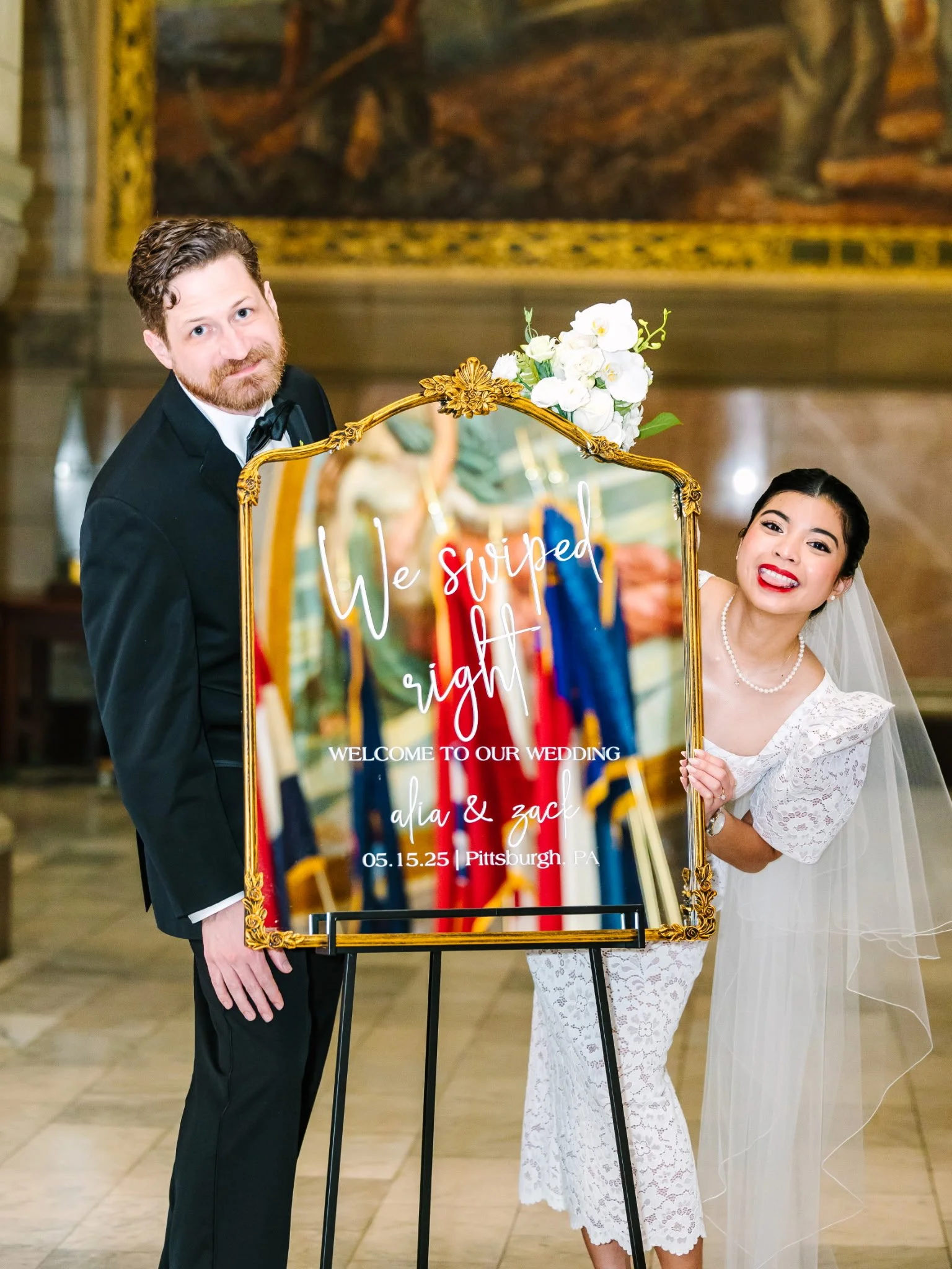 Wedding couple posing behind a welcome sign with a gold frame, flowers, and colorful ribbons, in an elegant hall.