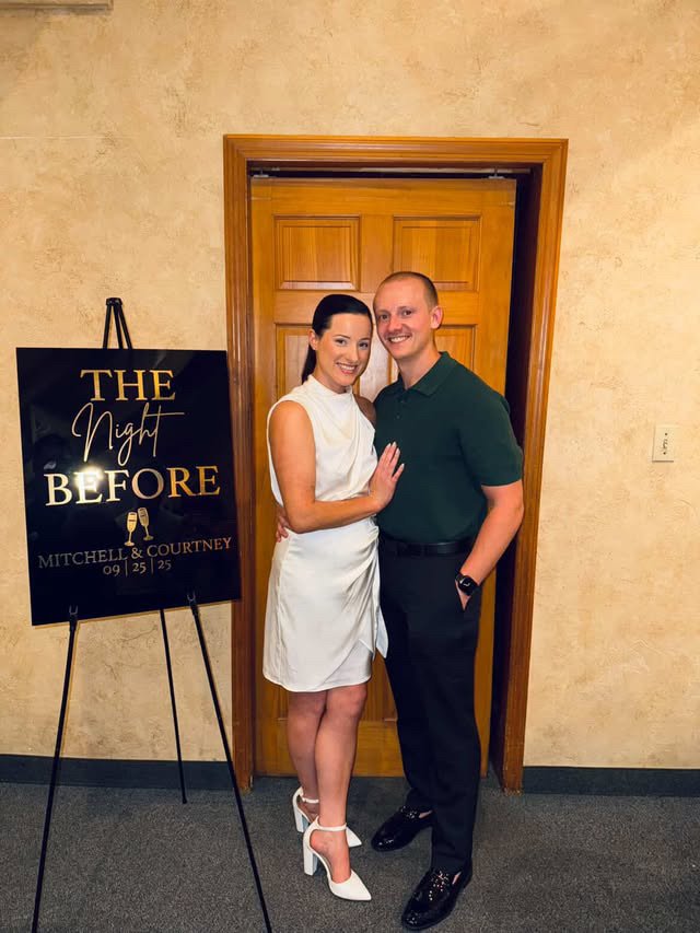 A smiling couple standing close together in front of a wooden door, with a sign on an easel that reads "The Night Before" and features a champagne glass graphic, indicating they are attending a wedding celebration for Mitchell and Courtney on Septemb