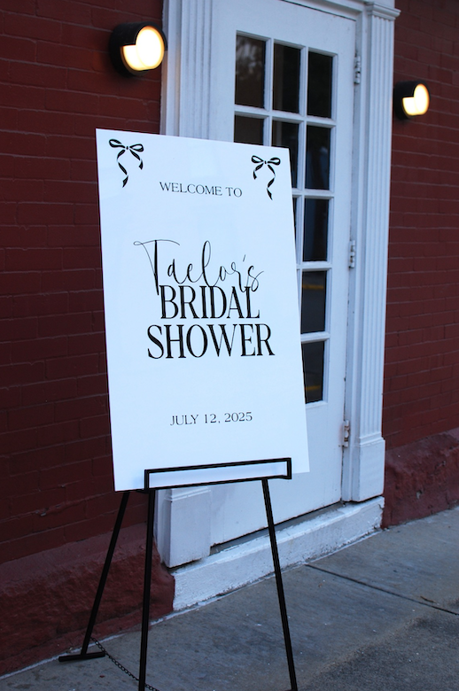 Sign for Taelor's bridal shower on July 12, 2025, outside a building with a red brick wall and a white door.