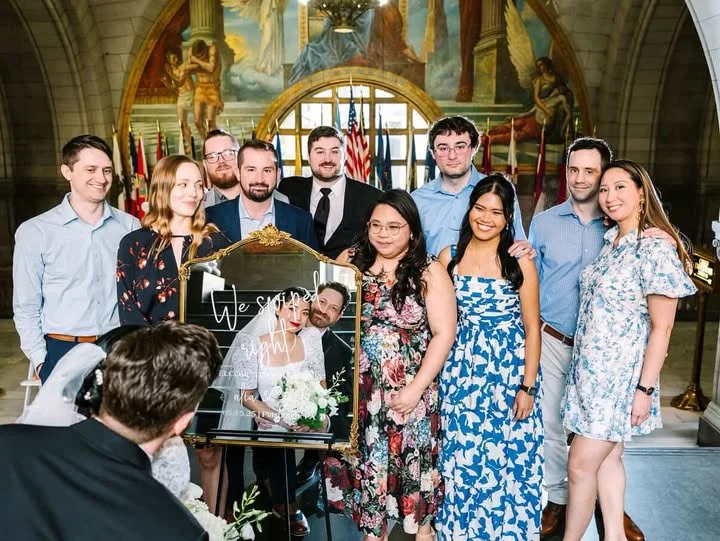 A wedding group photo inside a historic building with murals and flags in the background; ten people are smiling and gathered around a picture of the bride and groom.