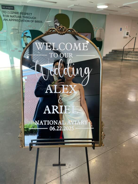 Mirror with wedding welcome sign for Alex and Ariel, dated June 22, 2025, featuring a photo of a bride in a white wedding dress and groom in a dark suit, located indoors with stairs and teal wall art in the background.