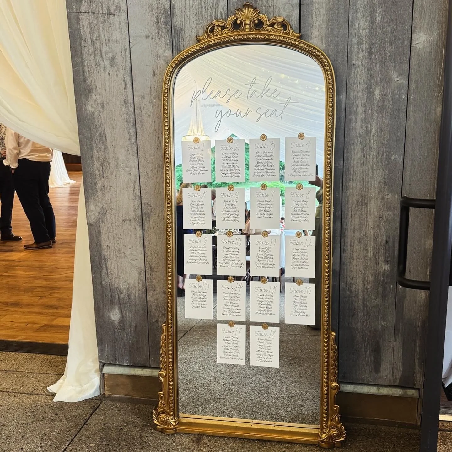 A large ornate full-length mirror on a wooden wall with a seating chart for a wedding or event reflected in it. The top of the mirror has a handwritten sign that says 'please take your seat'. Several pages with table assignments are taped to the mirr