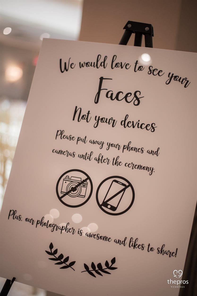 Sign at a wedding asking guests to see faces not devices and to put away phones and cameras. Includes icons of a camera and a phone with a line through them, and a message about the photographer sharing photos.