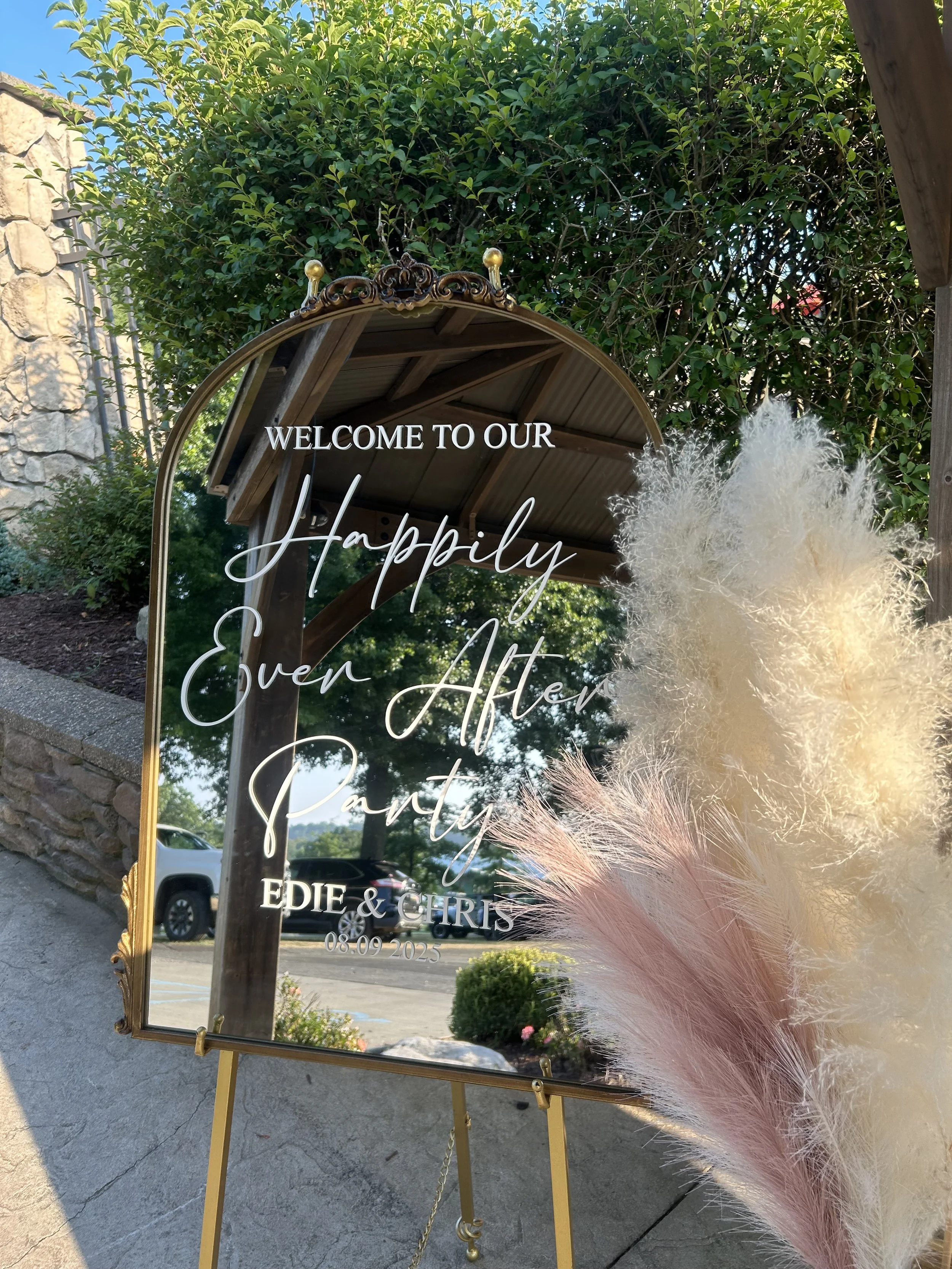 Mirror with a gold ornate frame displaying a welcome message for a celebration, decorated with pink and white pampas grass, with trees, cars, and a stone wall in the background.