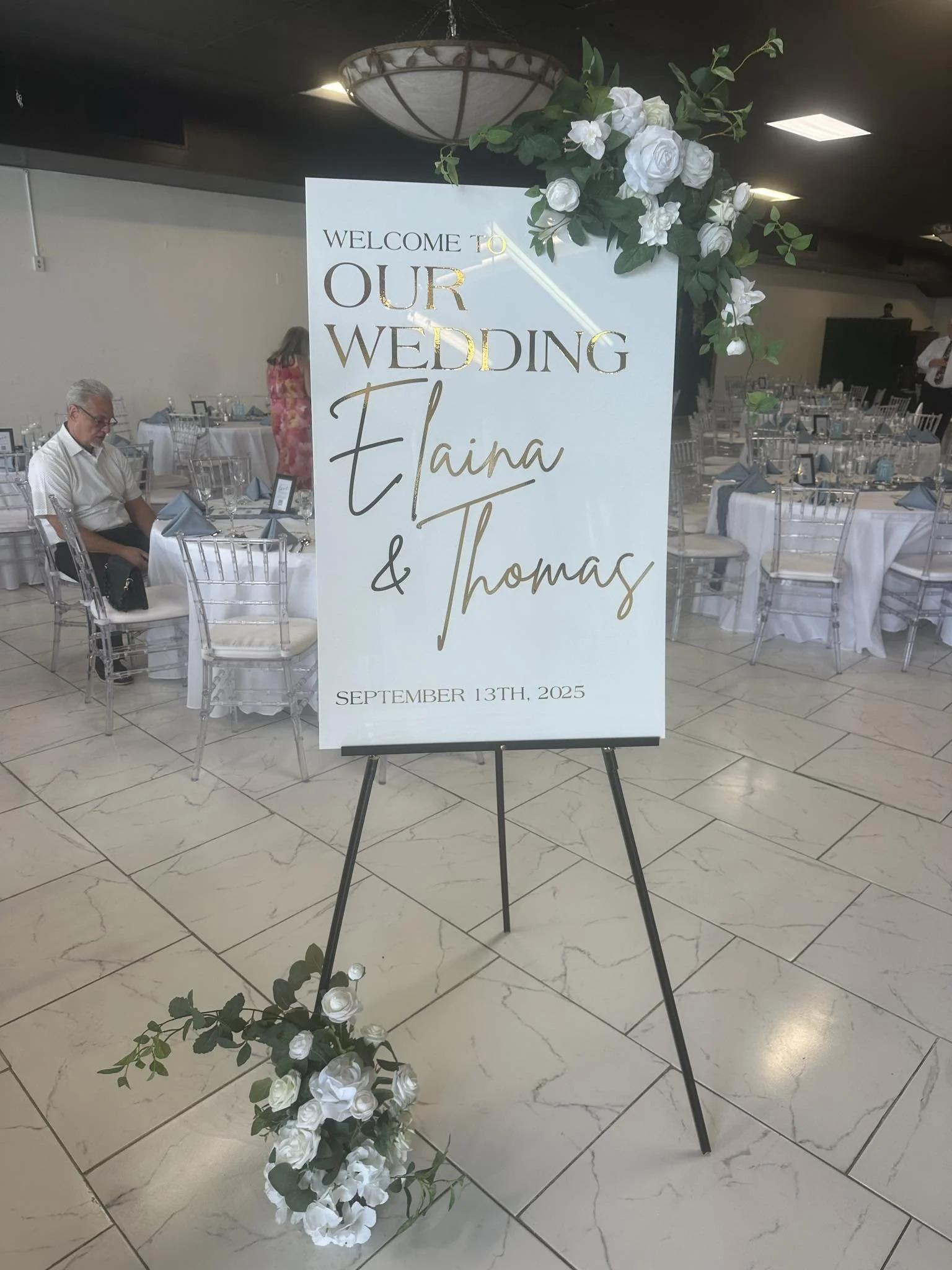 Wedding welcome sign with floral decorations, announcing the marriage of Elaina and Thomas on September 13th, 2025, placed in a decorated reception hall.
