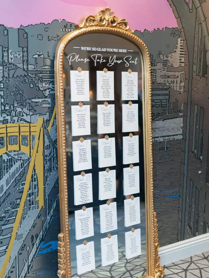 A decorative wedding seating chart displayed on a tall, ornate gold-framed mirror with handwritten instructions and guest lists on individual cards, set against a mural of a cityscape background.
Margaret James Photographer