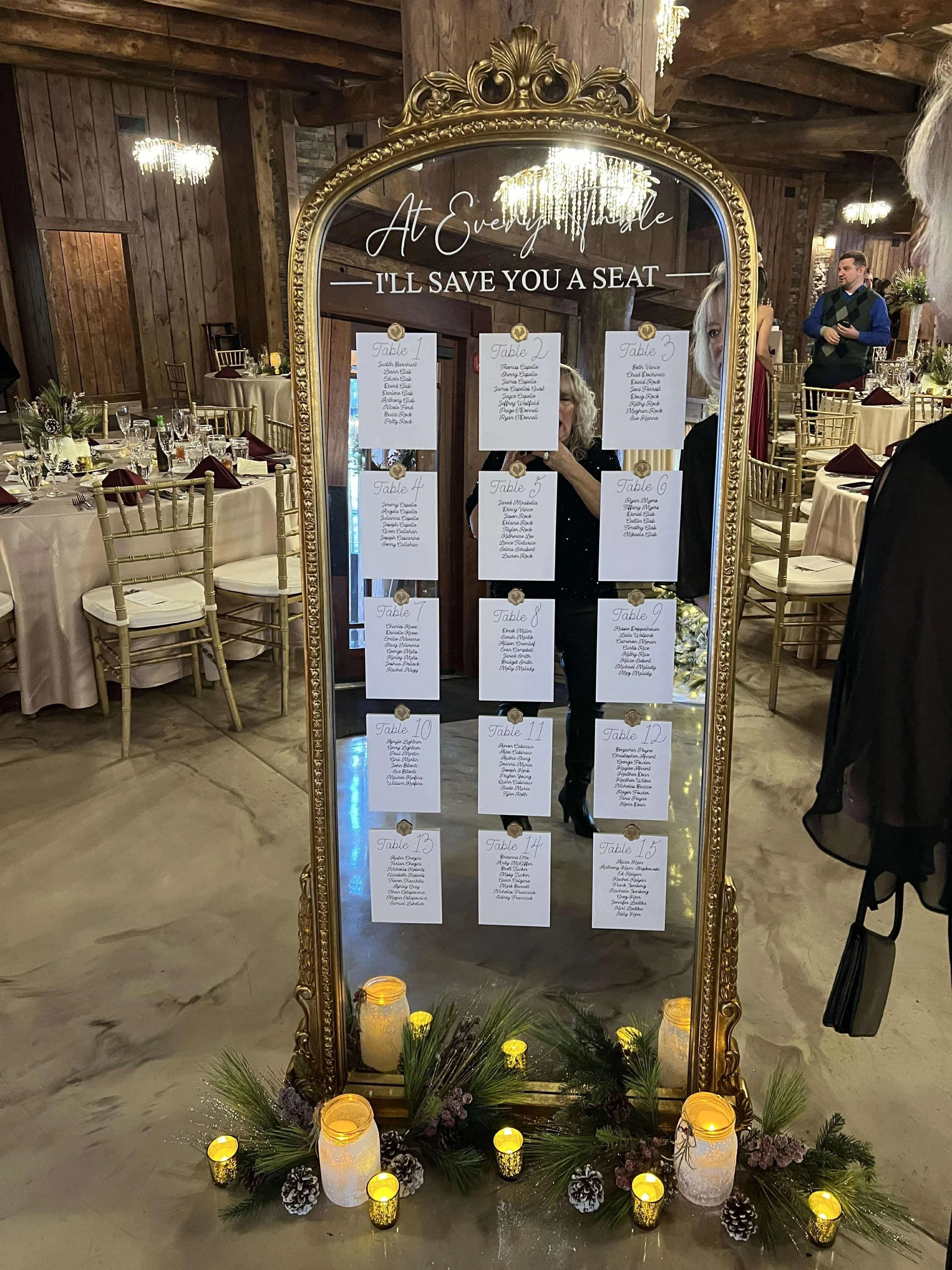 Decorative gold mirror with seating chart for a wedding, reading 'At Every Mile I’ll Save You a Seat,' decorated with candles, pine branches, pinecones, and flowers at the base, and illuminated chandeliers in the background.