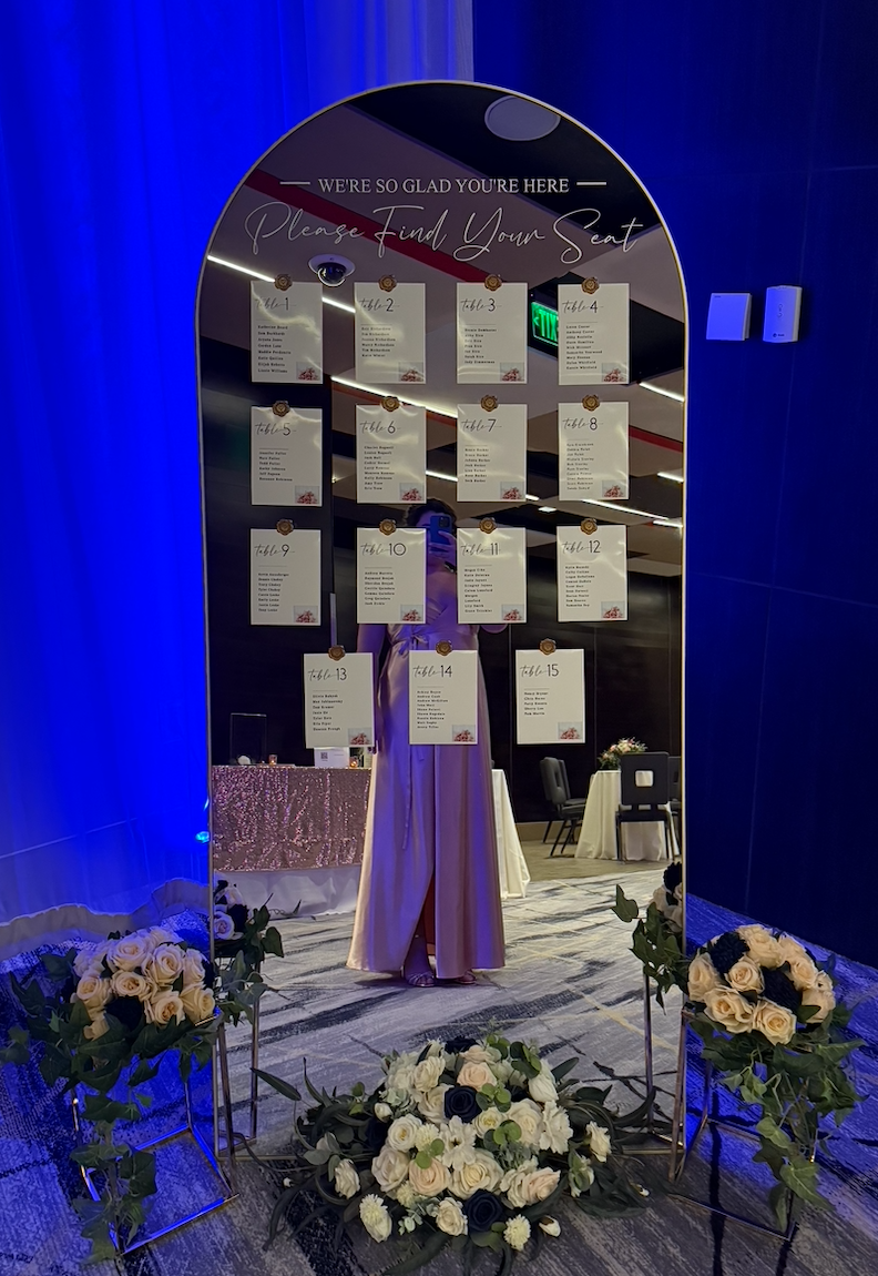 A decorative mirror with a sign at the top that reads "We're so glad you're here, Please Find Your Seat" and is surrounded by floral arrangements and seating charts with numbered tables inside a banquet hall.