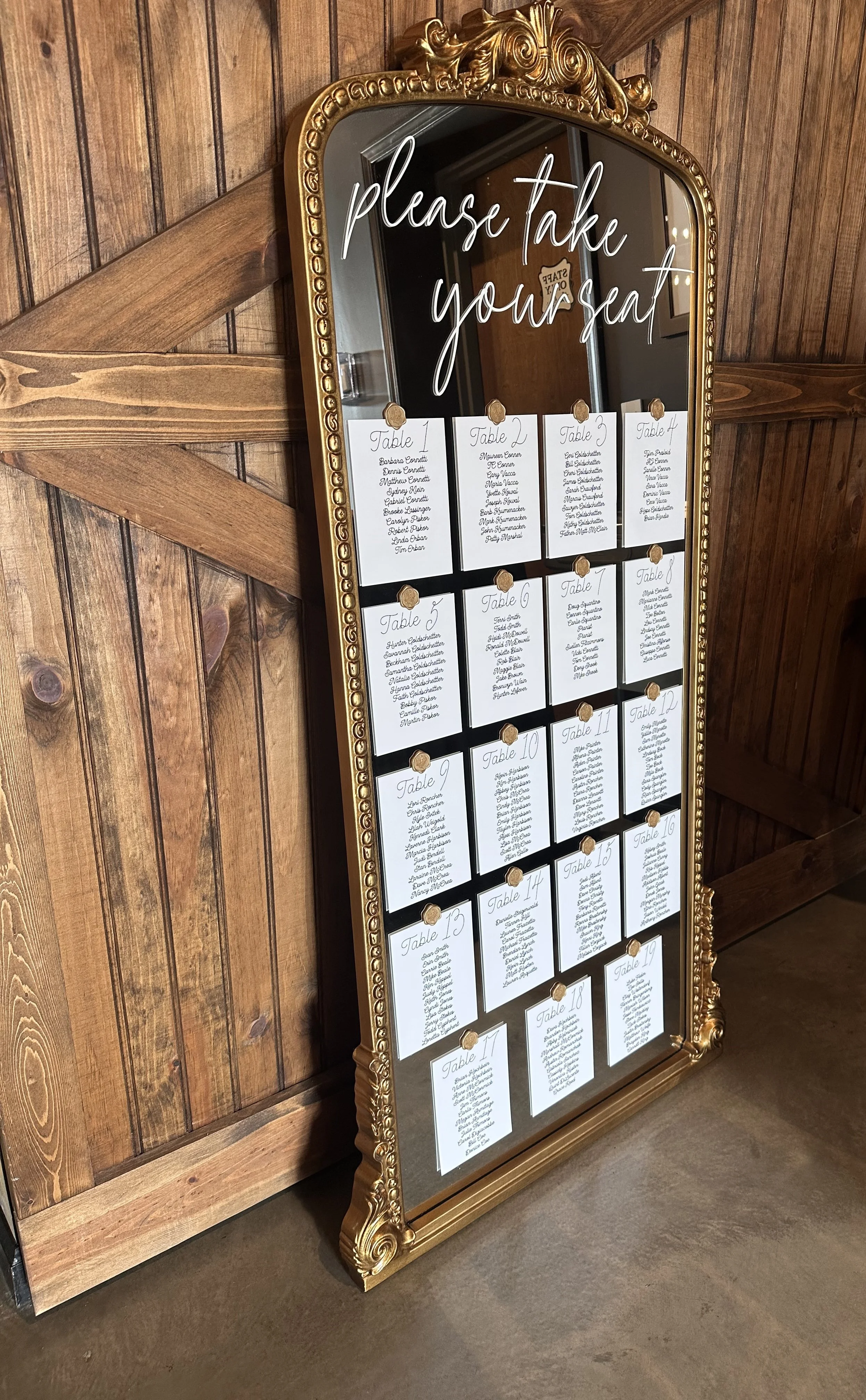 Mirror with gold ornate frame displaying table assignments for an event, with the phrase 'please take your seat' written on the glass and numbered tables with guest names on paper cards pinned to the mirror.
