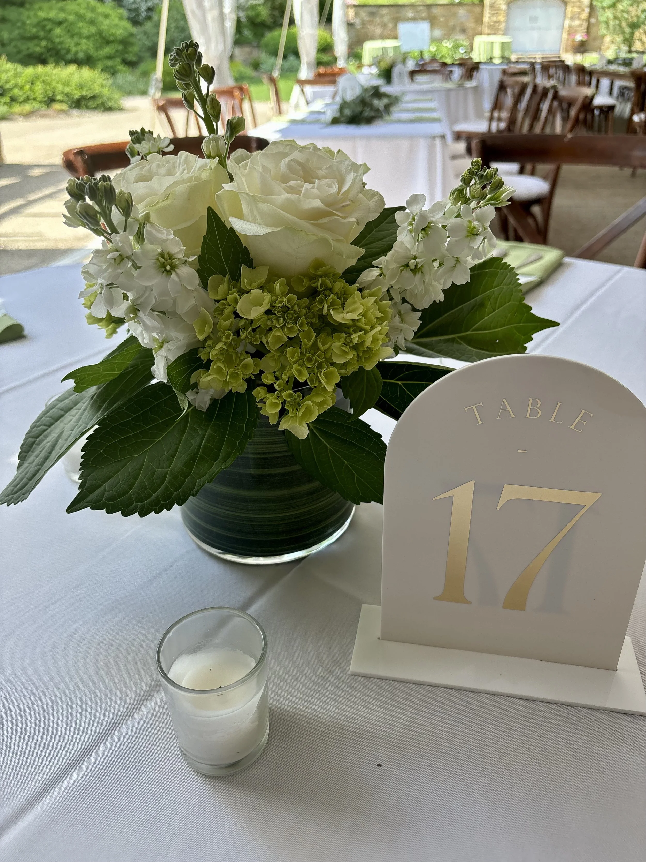 A white floral centerpiece with roses and hydrangeas in a dark ceramic vase, a table number sign with the number 17, and a small white candle in a glass holder on a white tablecloth at an outdoor event.