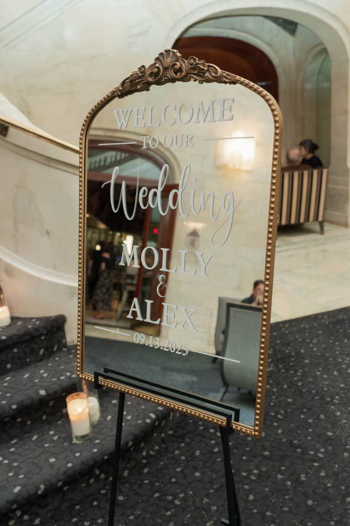 Decorative wedding welcome sign with ornate gold frame, reading "Welcome to our wedding Molly & Alex 09.13.2023" at a hotel or banquet hall entrance, with candles on the steps nearby.