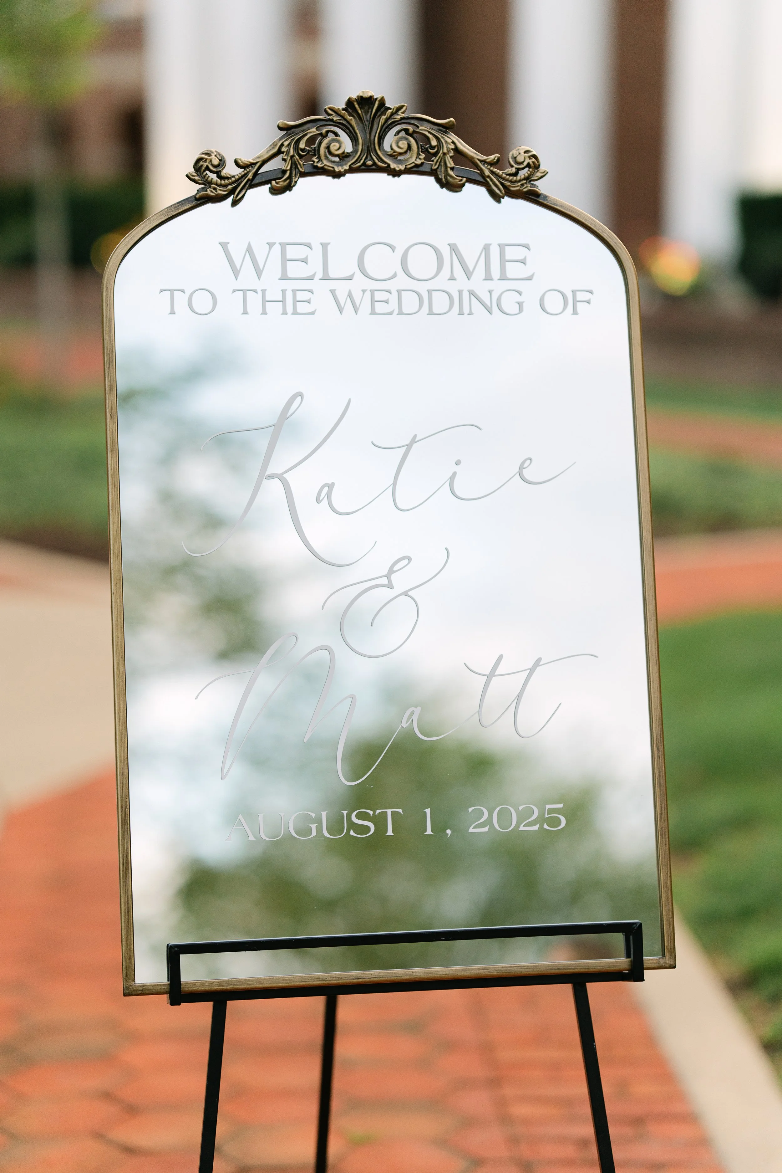 A decorative wedding sign with a gold ornate top, welcoming guests to the wedding of Kate and Matt on August 1, 2025, placed on an easel outdoors.

Kelli Slavonic Photographer